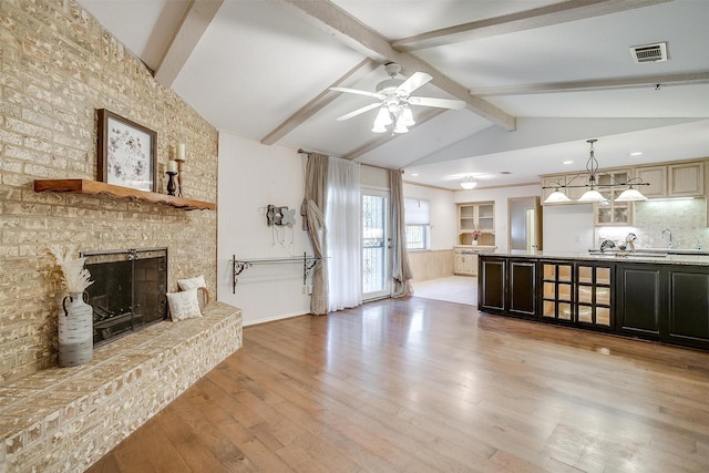 unfurnished living room with light wood finished floors, visible vents, a brick fireplace, vaulted ceiling with beams, and ceiling fan