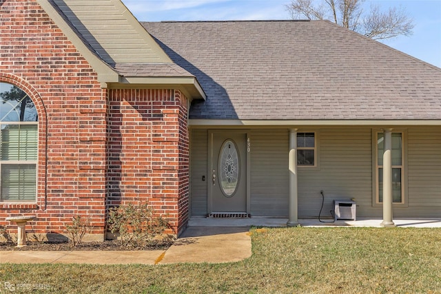 view of exterior entry featuring a yard, central AC unit, brick siding, and roof with shingles