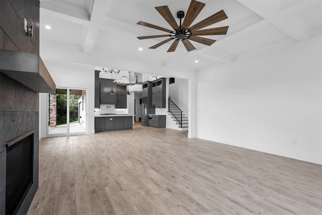 unfurnished living room featuring a tiled fireplace, ceiling fan with notable chandelier, coffered ceiling, stairway, and light wood-style floors