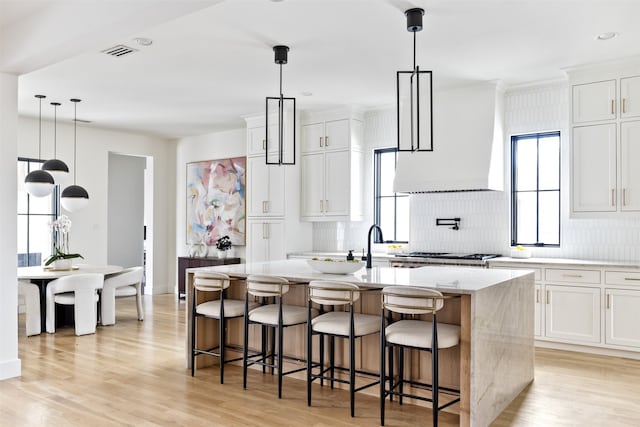 kitchen featuring an island with sink, custom range hood, light stone counters, tasteful backsplash, and light wood finished floors