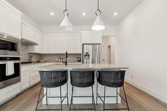 kitchen with under cabinet range hood, light wood-type flooring, a kitchen bar, decorative backsplash, and appliances with stainless steel finishes