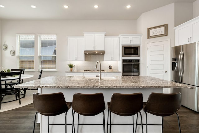 kitchen with dark wood-type flooring, a center island with sink, under cabinet range hood, stainless steel appliances, and decorative backsplash