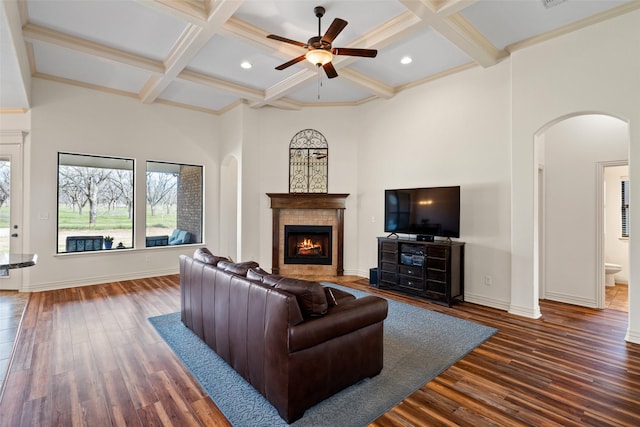 living area with dark wood-type flooring, a tiled fireplace, coffered ceiling, arched walkways, and ceiling fan