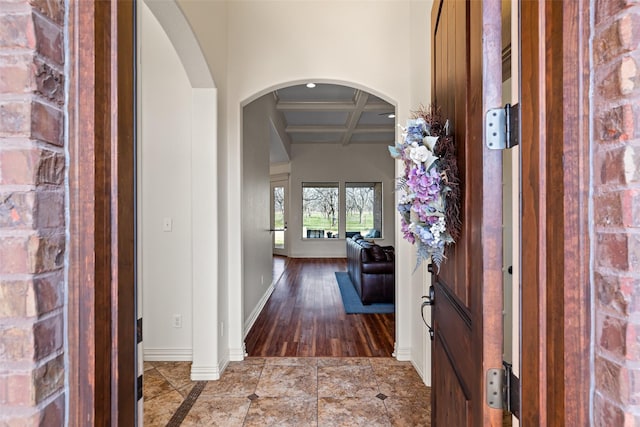 hallway featuring wood finished floors, baseboards, coffered ceiling, beam ceiling, and arched walkways