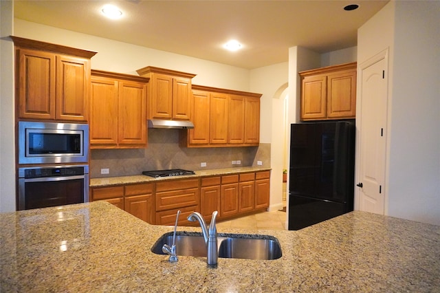 kitchen featuring light stone countertops, a sink, decorative backsplash, black appliances, and under cabinet range hood