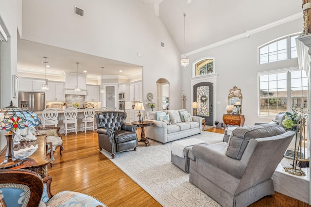 living room featuring light wood finished floors, visible vents, crown molding, arched walkways, and high vaulted ceiling