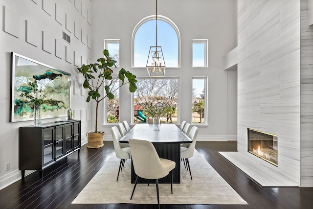 dining room featuring baseboards, dark wood finished floors, a premium fireplace, a towering ceiling, and an inviting chandelier