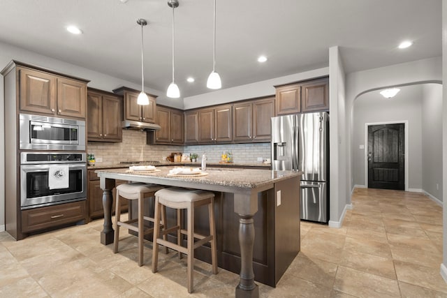 kitchen featuring a center island with sink, tasteful backsplash, stainless steel appliances, light stone countertops, and hanging light fixtures