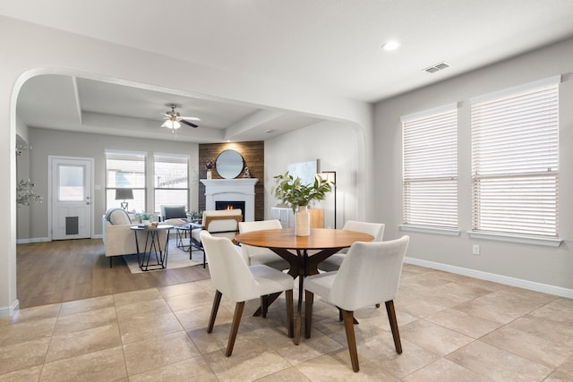 dining space featuring visible vents, baseboards, a lit fireplace, a raised ceiling, and a ceiling fan