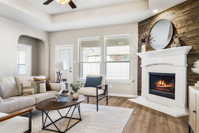 living area with a ceiling fan, arched walkways, a fireplace, light wood finished floors, and a raised ceiling