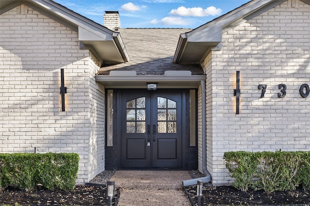 entrance to property with brick siding, french doors, a chimney, and a shingled roof