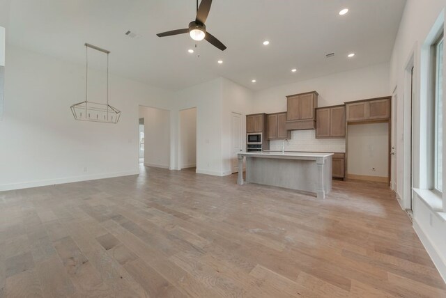 kitchen featuring backsplash, open floor plan, a center island with sink, light wood-style flooring, and a ceiling fan