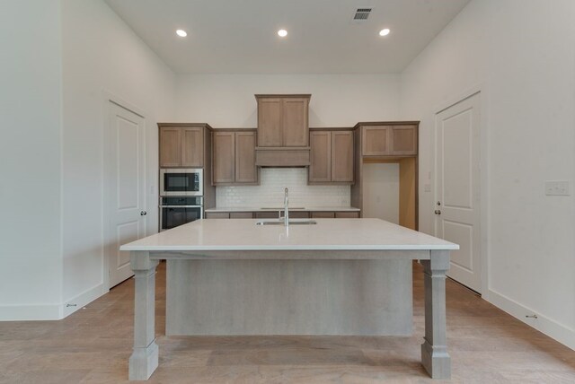 kitchen featuring oven, a kitchen island with sink, a sink, stainless steel microwave, and tasteful backsplash