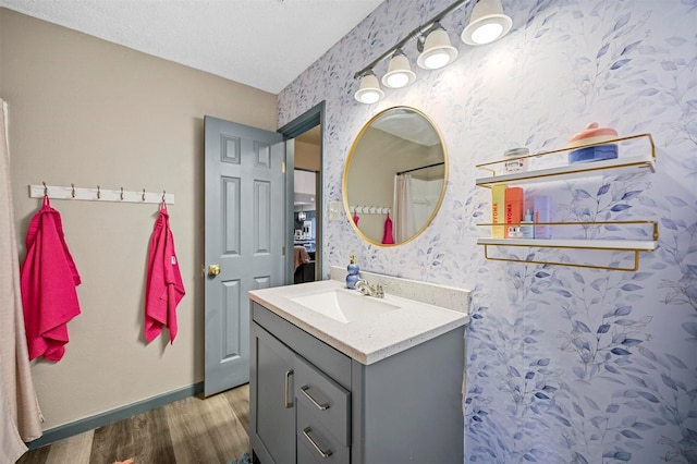 bathroom with a textured ceiling, wood finished floors, wallpapered walls, baseboards, and vanity