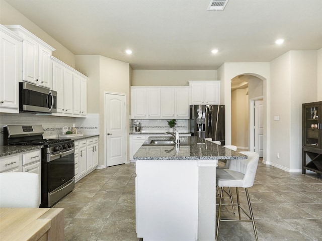 kitchen with an island with sink, arched walkways, a sink, appliances with stainless steel finishes, and backsplash