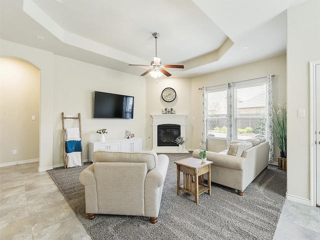 living area featuring a ceiling fan, baseboards, arched walkways, a glass covered fireplace, and a raised ceiling