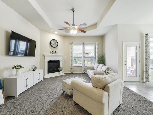 living area featuring baseboards, a tray ceiling, light tile patterned floors, a glass covered fireplace, and a ceiling fan