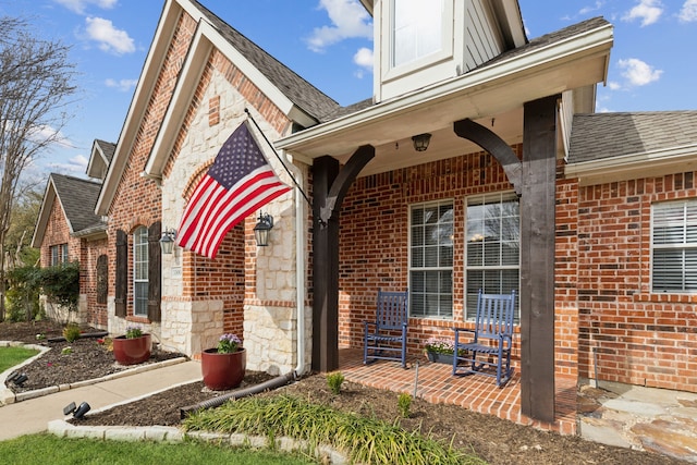 doorway to property featuring stone siding, brick siding, a porch, and roof with shingles