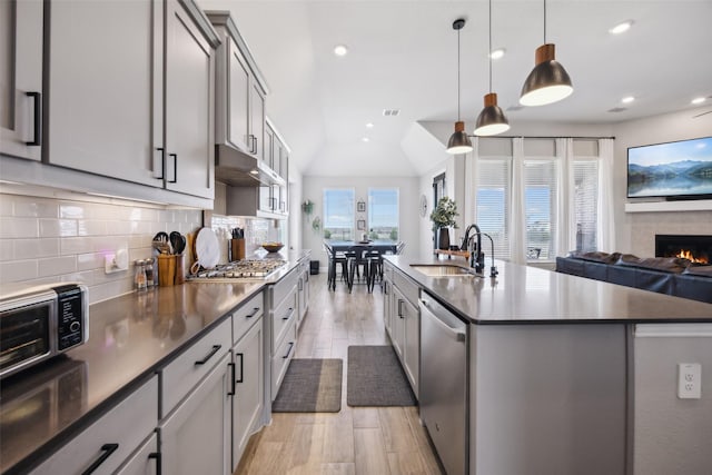 kitchen featuring a sink, under cabinet range hood, backsplash, a warm lit fireplace, and appliances with stainless steel finishes