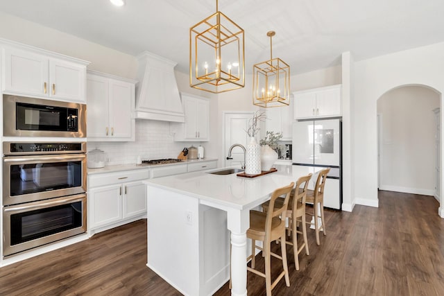 kitchen featuring custom range hood, decorative backsplash, arched walkways, stainless steel appliances, and a sink