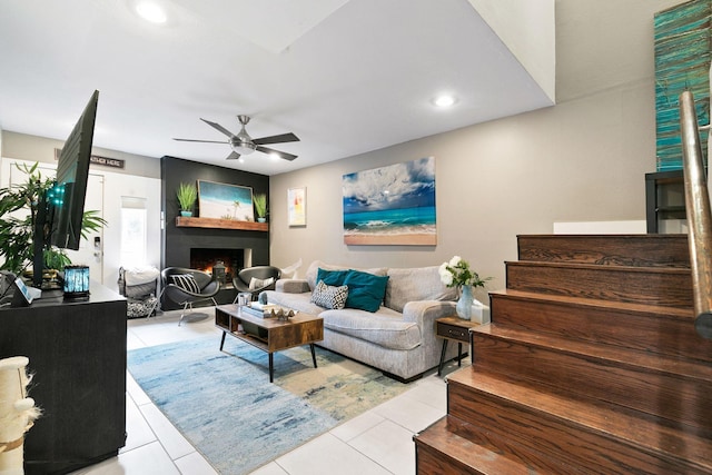 living room with stairs, light tile patterned floors, a warm lit fireplace, and ceiling fan