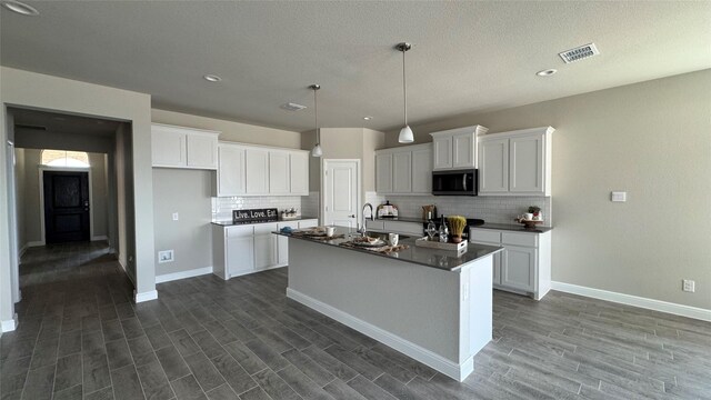 kitchen with dark wood finished floors, a kitchen island with sink, decorative backsplash, white cabinets, and dark countertops