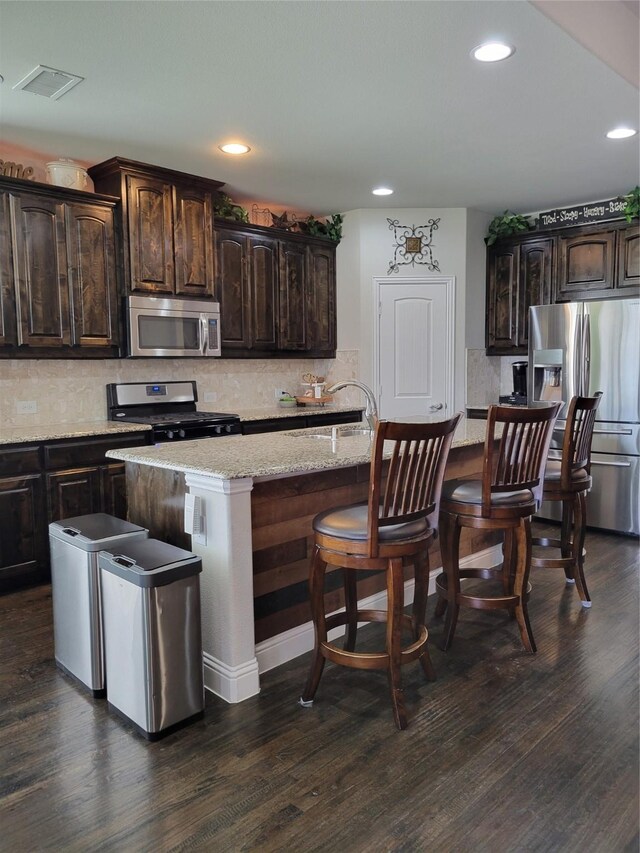 kitchen featuring dark brown cabinetry, a center island with sink, a kitchen bar, backsplash, and appliances with stainless steel finishes