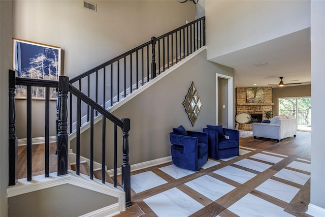staircase featuring inlaid floor details, baseboards, a brick fireplace, ceiling fan, and a high ceiling