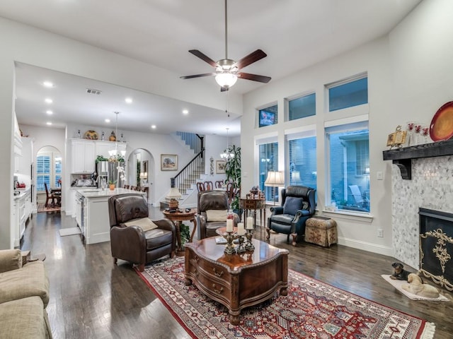 living room with dark wood-type flooring, stairway, a stone fireplace, ceiling fan with notable chandelier, and arched walkways