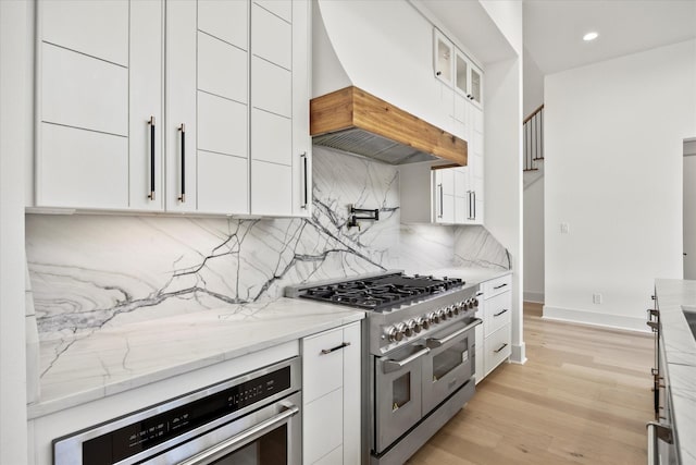 kitchen with light stone countertops, double oven range, white cabinets, premium range hood, and backsplash