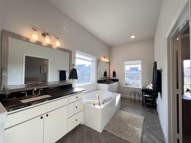 bathroom with a garden tub, two vanities, a healthy amount of sunlight, and a sink