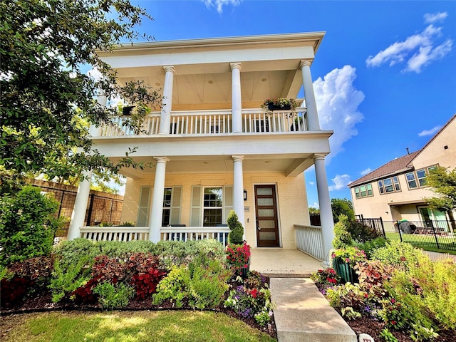 greek revival inspired property with a balcony, covered porch, brick siding, and fence