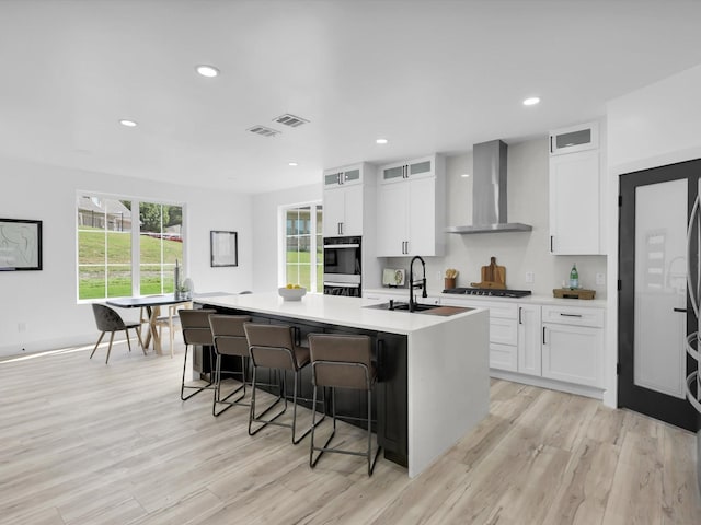 310 Harborview Dr, Rockwall, TX 75032 - kitchen featuring wall chimney range hood, a sink, a kitchen island with sink, white cabinets, and light wood-style floors