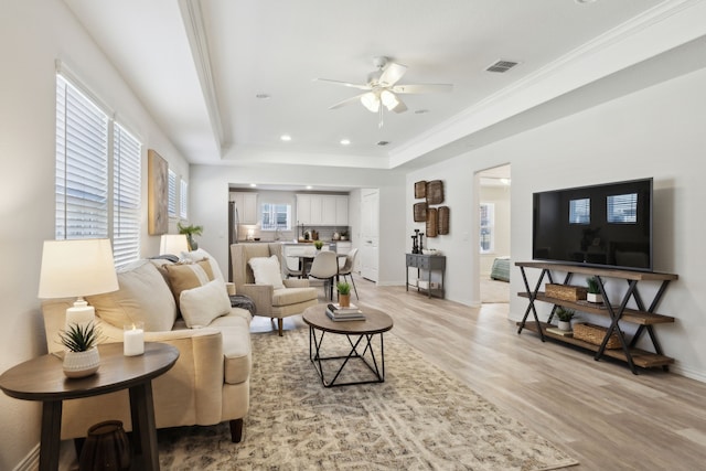 living area with light wood finished floors, a raised ceiling, visible vents, baseboards, and ceiling fan