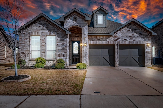 french country inspired facade with stone siding, concrete driveway, an attached garage, a front yard, and brick siding