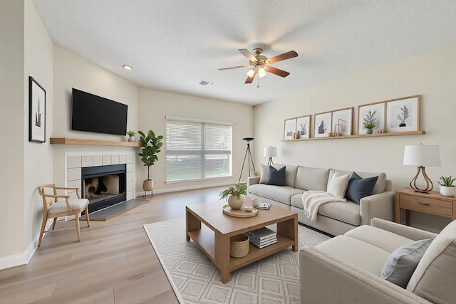 living room featuring visible vents, a tiled fireplace, light wood-style floors, a textured ceiling, and a ceiling fan