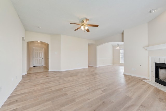 unfurnished living room featuring arched walkways, a tile fireplace, light wood-style floors, and a ceiling fan