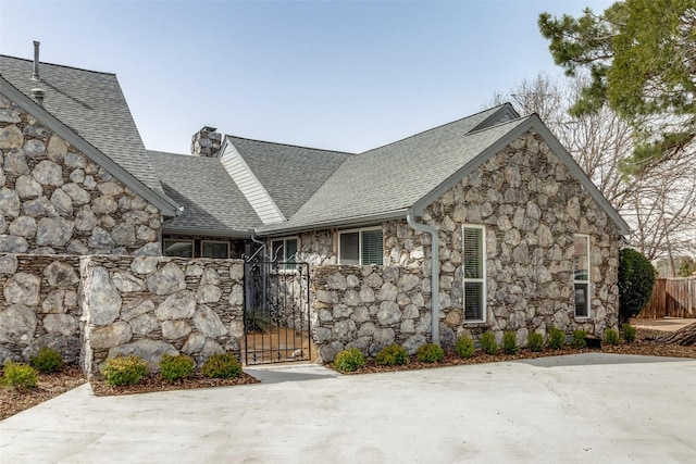 view of front facade featuring a shingled roof, stone siding, a chimney, fence, and a gate