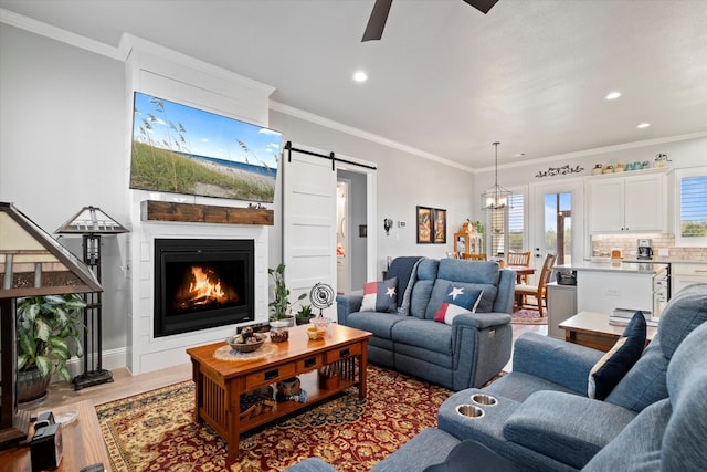 living room with wood finished floors, ornamental molding, a barn door, ceiling fan with notable chandelier, and a large fireplace
