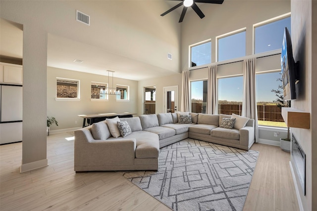 living area with visible vents, light wood finished floors, baseboards, a towering ceiling, and ceiling fan with notable chandelier