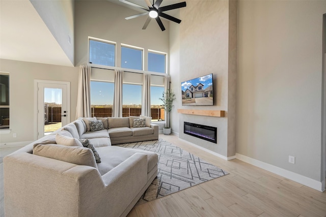 living area featuring a glass covered fireplace, baseboards, a towering ceiling, wood finished floors, and ceiling fan