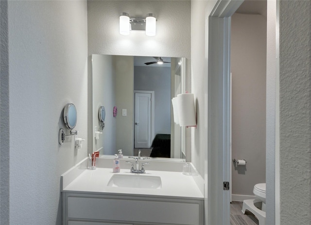 bathroom featuring ceiling fan, baseboards, toilet, a textured wall, and vanity