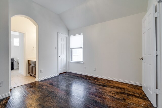 unfurnished bedroom featuring baseboards, vaulted ceiling, ensuite bath, arched walkways, and wood finished floors