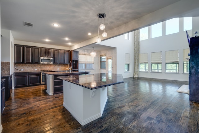 kitchen featuring stainless steel microwave, dark wood-style floors, dark brown cabinets, a center island with sink, and visible vents