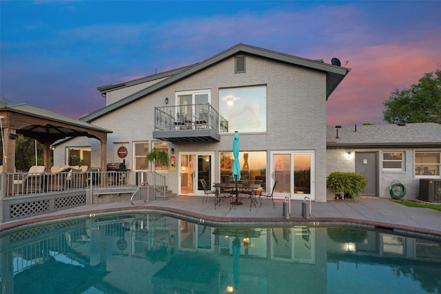 back of house featuring central air condition unit, a patio area, brick siding, an outdoor pool, and a balcony