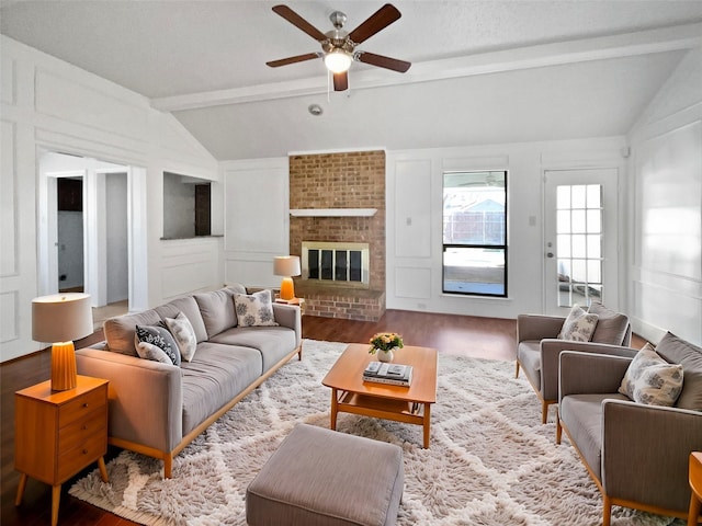 living room with wood finished floors, a ceiling fan, vaulted ceiling with beams, a brick fireplace, and a decorative wall