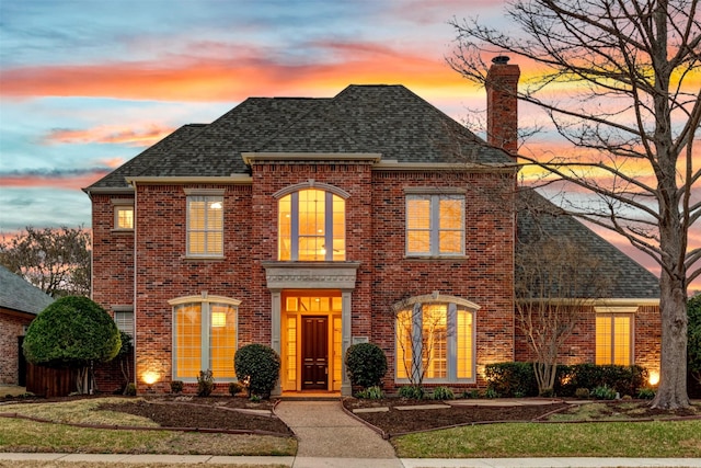 view of front of property with brick siding, roof with shingles, a chimney, and a lawn