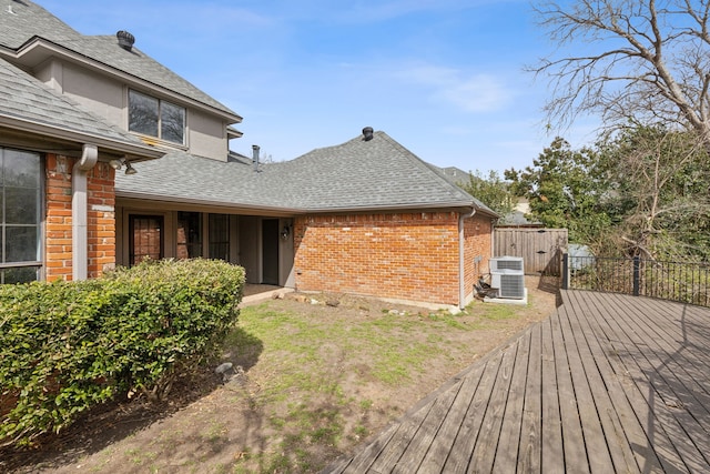 view of side of property with a deck, fence, a shingled roof, brick siding, and central AC unit