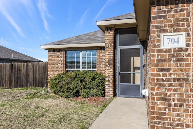 property entrance featuring fence, a shingled roof, brick siding, and a yard