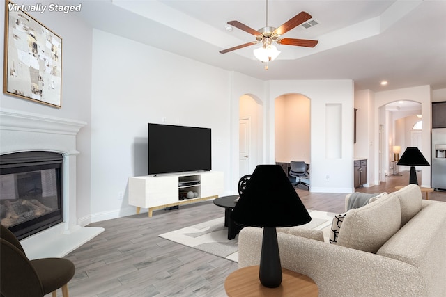 living area with arched walkways, a tray ceiling, a ceiling fan, and light wood-type flooring